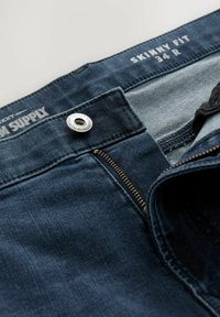 Úzke deniminové džínsy v tmavomodrej farbe, s strieborným cvokom a zipsom, s svetlejšou látkou vo vnútri pásu a vrecka.