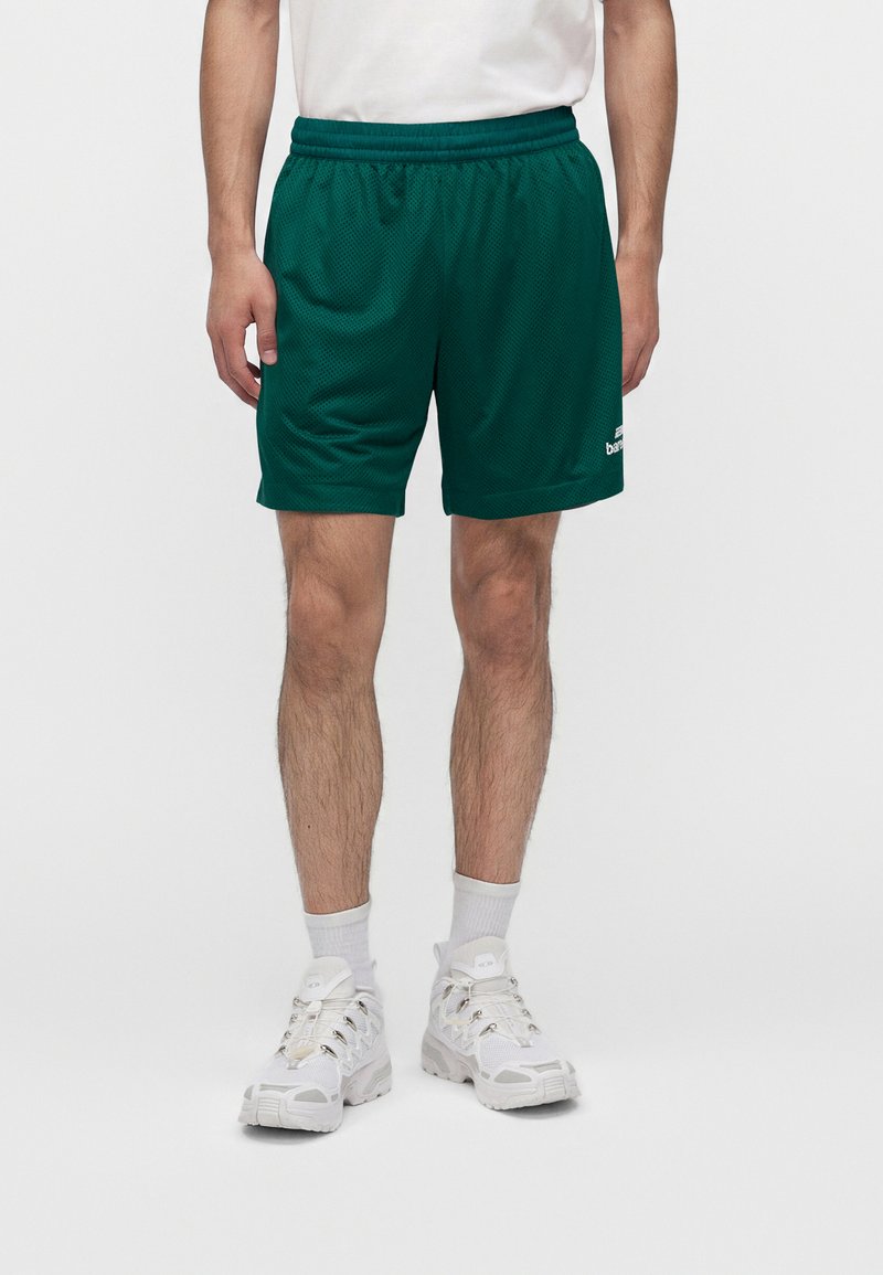 Homme portant un short de sport en mesh vert foncé, des chaussettes blanches et des baskets blanches épaisses, debout devant un fond uni.