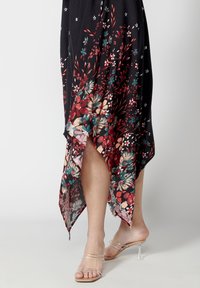 Jambes dans des sandales à talons hauts transparentes à brides, portant une jupe noire avec un motif floral rouge, bleu sarcelle et crème et un ourlet asymétrique.
