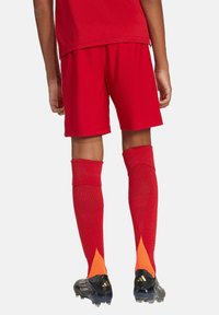 Rode sportieve shorts met bijpassende rode kniekousen met een oranje patroon aan de bovenkant. Zwarte voetbalschoenen met drie strepen.