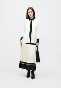 Biela plisovaná blúzka s čiernymi akcentmi, doplnená béžovou plisovanou sukňou s tmavým lemom. Modelka drží čiernu kabelku a nosí čierne čižmy.