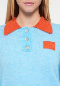 Gestrickter Pullover in Hellblau mit orangefarbenem Kragen, drei Knöpfen und einem kleinen orangefarbenen Fleck auf der linken Seite. Weiche Textur.