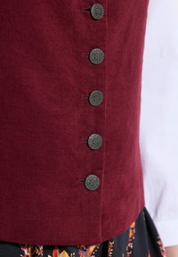 Burgundy corduroy-vesti, jossa on kuusi koristeellista tummaa nappia, sekä ribattua tekstuuria ja muototettua siluettia.