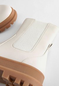 Bottines blanches imperméables avec un panneau arrière texturé, semelle en caoutchouc et une languette de tirage, présentant une finition lisse et des accents nervurés.