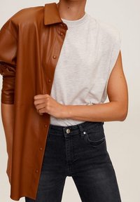 Braune Hemdjacke aus Kunstleder mit Druckknöpfen über einem grauen T-Shirt. Schwarze, hoch taillierte Jeans mit Vorderknopf und Taschendetail.