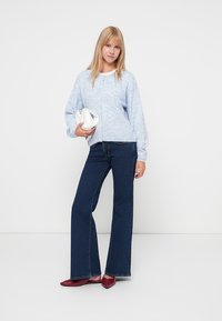 Lyseblå, struktureret, langærmet sweater med rund halsudskæring, parret med mørkeblå flagrende jeans og røde slip-on sko. Holder en hvid taske.