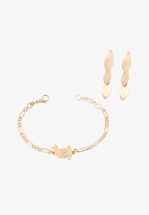 Kultainen rannekoru pupu-riipuksella ja rusetilla, yhdistettynä kultaiset kierretyt roikkuvat korvakorut valkoisella taustalla.