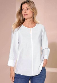 Camicia bianca con scollatura tonda, maniche lunghe e dettaglio con bottone a chiave sul davanti. Orlo leggermente curvo e texture del tessuto liscia.