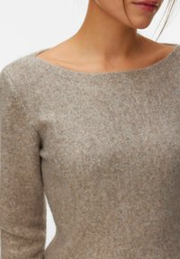 Femme portant un pull en maille beige doux avec un col large et arrondi et des manches longues, vue de l'épaule au milieu du torse.