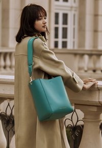 Borsa a secchiello in pelle verde acqua con tracolla abbinata rimovibile, texture liscia, dettaglio logo discreto e forma strutturata. Posizionata su uno sfondo neutro.
