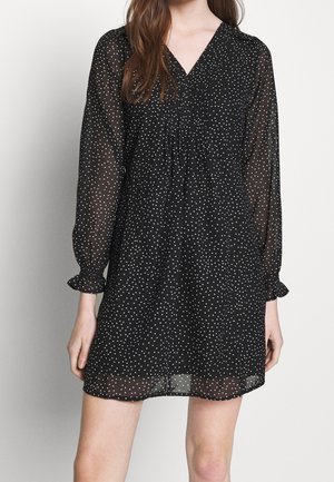 Robe noire semi-transparente à pois blancs, col en V, manches longues avec poignets froncés, et coupe décontractée.