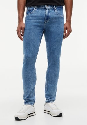 Άνδρας που φορά ανοιχτό μπλε στενά τζιν και λευκά αθλητικά παπούτσια, στέκεται μπροστά σε απλό λευκό φόντο.