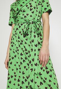 Robe portefeuille verte à manches courtes avec des motifs de cœurs noirs, nouée à la taille avec une ceinture en tissu, portée par une personne avec les bras et la main visibles.