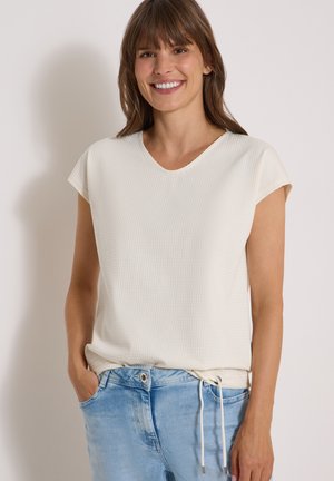 Mujer sonriente con cabello castaño que lleva una camiseta blanca de manga corta y vaqueros azul claro, de pie con una mano en el bolsillo.