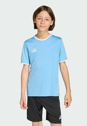 Młoda osoba ubrana w jasnoniebieską sportową koszulkę i czarne szorty z logo Adidas, stojąca na jednolitym jasnym tle.