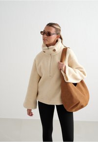 Krémový fleece pulovr s vysokým límcem a dvěma hnědými knoflíky, doplněný béžovou semišovou taškou a černými legínami.