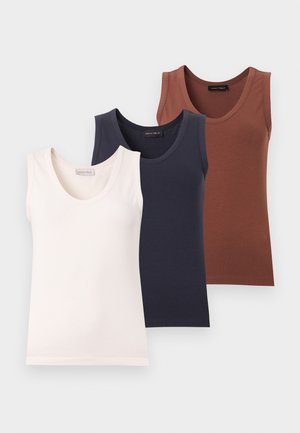 Femme portant des lunettes de soleil noires, un top blanc sans manches, un jean évasé bleu et des baskets blanches, debout devant un fond blanc uni.