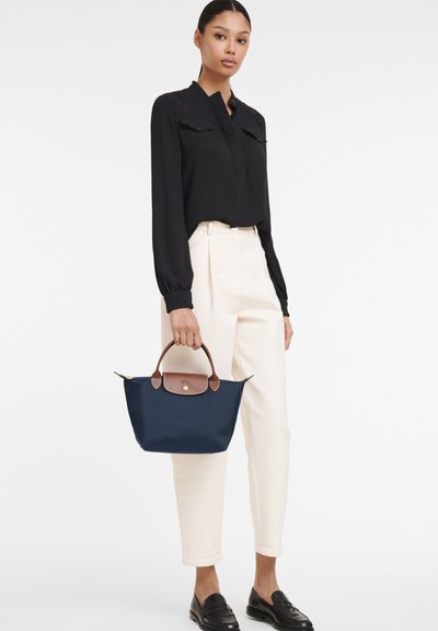 Marineblaue Stoffhandtasche mit braunen Lederakzenten, mit einem runden Griff und einem Reißverschluss, gehalten von einer Person in einer schwarzen Bluse und cremefarbenen Hosen.
