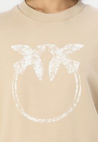 Beige színű póló kerek nyakkivágással, amelyen a fronton két madár és egy kör alakú keret texturált fehér grafikája látható.