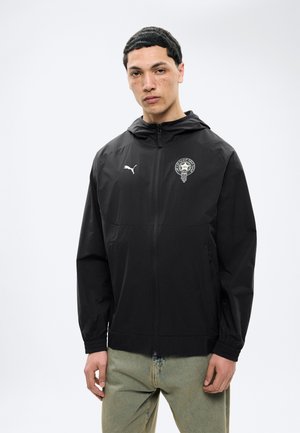 Noor mees lühikeste mustade juustega, kes kannab musta Puma tõmblukuga jopet, millel on logo ja embleem, seisab ühevärvilise valge tausta ees.