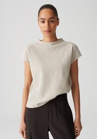 Light beige textured top with short sleeves, round neckline, and relaxed fit; pairs with dark pleated trousers for contrast.