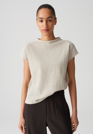 Light beige textured top with short sleeves, round neckline, and relaxed fit; pairs with dark pleated trousers for contrast.