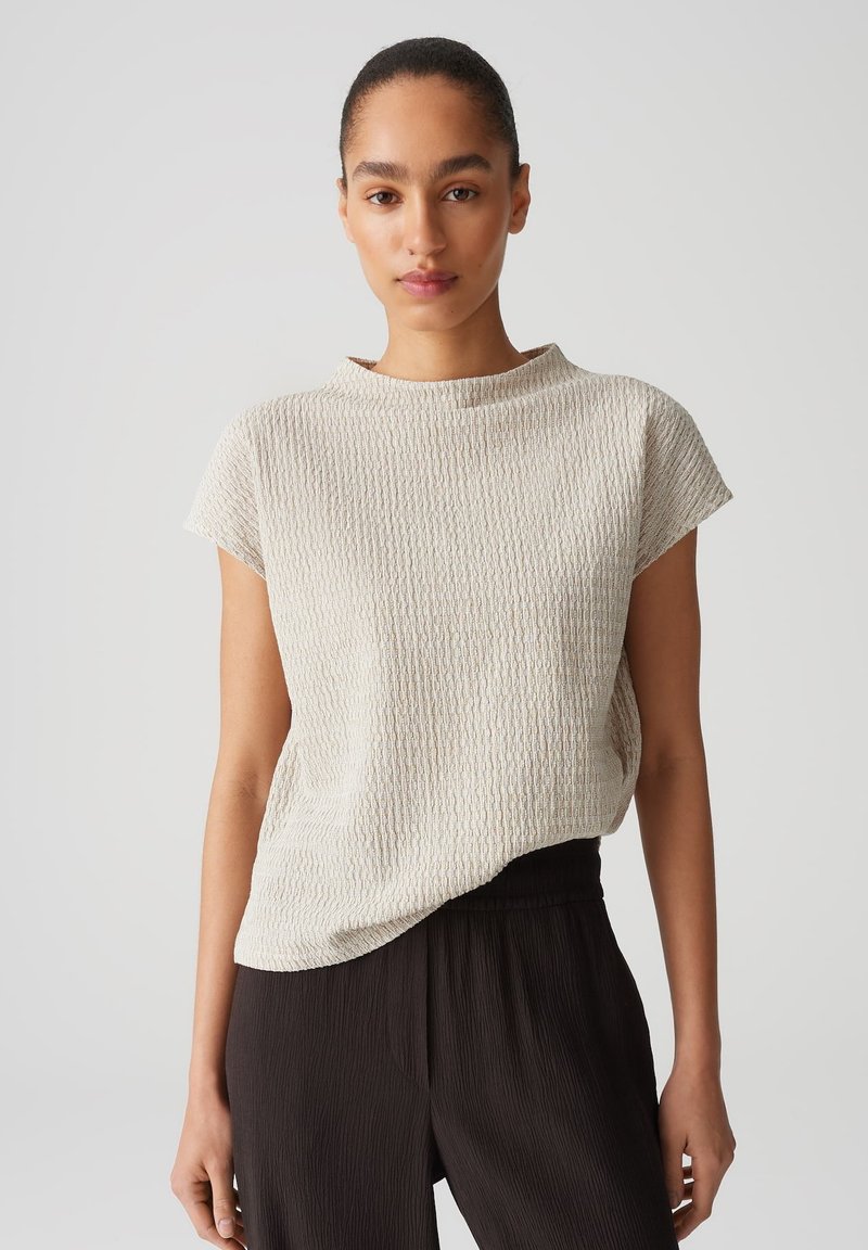 Light beige textured top with short sleeves, round neckline, and relaxed fit; pairs with dark pleated trousers for contrast.