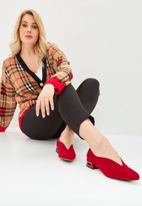 Rote Wildleder-Pumps mit einem tiefen V-Ausschnitt und einem klobigen Absatz. Getragen mit schwarzen, schmal geschnittenen Hosen und einer gemusterten beigefarbenen Strickjacke.