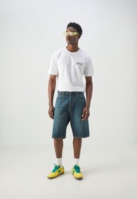 Witte T-shirt met logo, blauwe denim shorts en gele sneakers met groene accenten. Persoon draagt gele zonnebril en staat voor een lichte achtergrond.