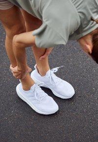 Personne portant des baskets blanches se penche et tient sa cheville, debout sur une surface asphaltée.