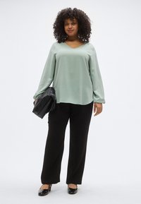 Μέντα πράσινη μπλούζα με μακριά μανίκια και V-λαιμόκοψη, σε συνδυασμό με μαύρο παντελόνι φαρδύ και λαμπερά μαύρα παπούτσια, κρατώντας μια μαύρη τσάντα φάκελο με υφή.