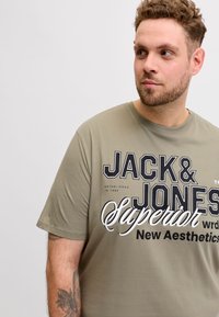 Άνδρας με σγουρά μαλλιά και γένια, φοράει πράσινη ελιά "Jack & Jones Superior" μπλούζα, κοιτάζει δεξιά μπροστά σε απλό φωτεινό φόντο.