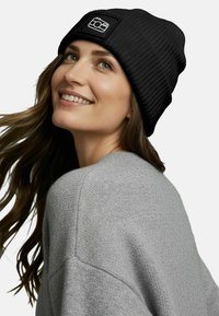 Black ribbed beanie with a textured finish, featuring a square logo patch. Worn with a gray sweater, showing soft fabric and a relaxed fit.