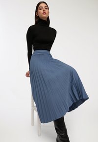 Una gonna a pieghe blu realizzata in tessuto morbido, a vita alta con una texture liscia, abbinata a un dolcevita nero a coste e stivali fino al ginocchio.