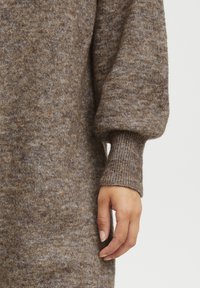 Manche de pull en tricot marron avec un motif marbré texturé. Détail de poignet côtelé complétant le design ample. Main placée près de l'ourlet.