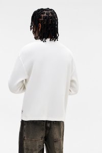 Fehér bordázott pulóver, hosszú ujjakkal, laza szabással és a szegélyén látható címkével, sötét, texturált nadrággal párosítva.