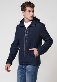 Chaqueta softshell azul marino con capucha, cierre de cremallera, dos bolsillos laterales y logotipo bordado en la parte frontal. Presenta una superficie suave y texturizada.