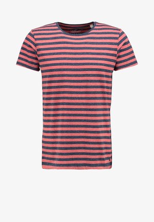 Młody mężczyzna w jasno-niebieskiej koszuli na czerwono-granatowej pasiastym t-shircie, z podwiniętymi jasnymi dżinsami i szarymi płóciennymi trampkami idący do przodu.