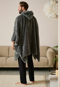 Texturiertes, dunkelgraues Kapuzenshirt mit weit geschnittenen Ärmeln und einem Saum, der zu einer Spitze herabhängt. Getragen über schwarzen Hosen, steht auf einem Teppich.