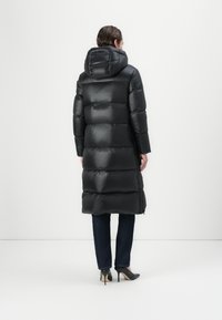 Zwarte gewatteerde lange jas met een capuchon, met een gladde, glanzende textuur en accentuerende naden. Gecombineerd met donkere jeans en hoge hakken.