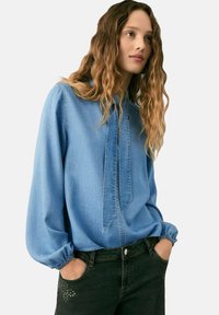 Camisa de mezclilla azul claro con cuello y mangas largas. Presenta un detalle de lazo en la parte delantera y puños elásticos. Combinada con vaqueros oscuros.