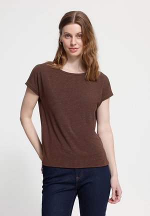 Mujer con el pelo largo castaño claro lleva una camiseta marrón de manga corta y vaqueros oscuros, de pie sobre un fondo blanco liso.