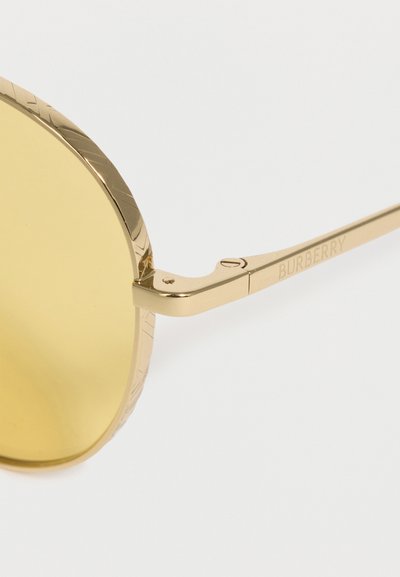 Στρογγυλά γυαλιά ηλίου με χρυσό τόνο, κίτρινους φακούς, χαραγμένη άκρη σκελετού και λογότυπο "BURBERRY" στον λεπτό βραχίονα.