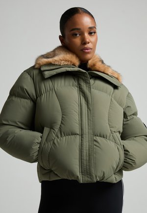 Femme portant une veste matelassée vert olive avec un col bordé de fourrure, les mains dans les poches, regardant la caméra avec confiance sur un fond uni.
