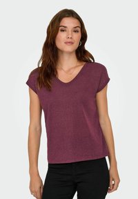 Femme aux longs cheveux bruns portant un haut maroon à manches courtes en V et un pantalon noir, se tenant contre un fond clair.