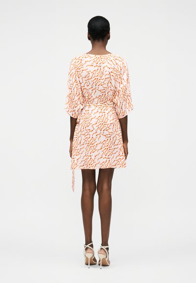 Γυναίκα που στέκεται με την πλάτη, φοράει κοντό λευκό φόρεμα με πορτοκαλί πουά σχέδιο και μπεζ ψηλοτάκουνα σανδάλια σε απλό φόντο.