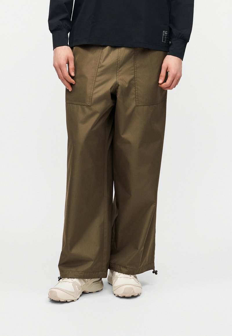 Άτομο που φορά χακί παντελόνι με φαρδιά πόδια και μπροστινές τσέπες, λευκά chunky αθλητικά παπούτσια, μαύρο μακρυμάνικο τοπ ορατό στη μέση.