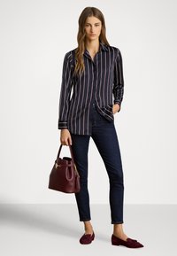 Sieviete tērpusies tumši zilsvītrainā blūzē un tumšās džinsās, tur rokā bordo krāsas somu un valkā saskaņotas bordo krāsas loafers kurpes.