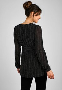 Svart, texturerad långärmad topp med silver vertikala ränder, en insvängd midja och en lätt peplumdesign. Har en rundad halsringning.