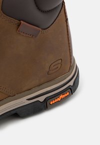 Brun læderstøvle med mørkegrå detaljer, gummisul, og orange "Goodstar" branding. Har stikninger og en tekstureret finish.